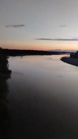 A video shows a view of a wide river at dusk. The sky is a soft gradient of colors, with hints of orange and pink near the horizon, transitioning to a pale blue and gray overhead. Thin, wispy clouds are scattered across the sky. The river itself is calm and reflective, mirroring the colors of the sky. The banks of the river are lined with dark silhouettes of trees and foliage on the left side, and a sandy bank with sparse vegetation on the right. The overall impression is one of peaceful twilight.