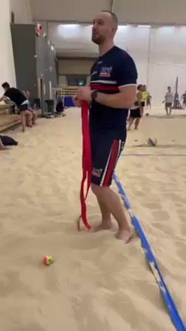 The video shows a man standing barefoot in a beach volleyball court. He is wearing a navy blue shirt with red and white stripes, navy blue shorts with red and white stripes, and a watch on his left wrist. He is holding a red ribbon. There is a blue boundary line in front of him and a small ball on the sand. In the background, there are other people playing beach volleyball and some people sitting on a bench. The scene is indoors.