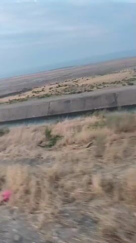 The video shows a view from a moving vehicle looking towards a large concrete canal with water flowing through it. The canal runs diagonally across the frame, disappearing into the distance. The surrounding landscape is dry and dusty, with sparse, brown vegetation. There are some green bushes growing along the edge of the canal. In the distance, there appear to be some structures or bridges over the canal, and possibly some people. The sky is overcast with a hazy, pale blue color. The camera is angled slightly downwards, and the motion of the vehicle creates a slight blur in the foreground.