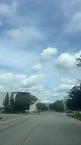 The video shows a street scene with industrial buildings and trees under a cloudy sky. A black SUV drives by on the road in the foreground. The building on the left has the number 130 visible on it.