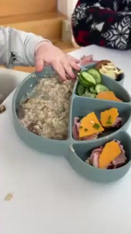 A child, wearing a gray long-sleeved shirt, reaches into a section of a teal silicone plate containing a light-colored, lumpy mixture of food. The plate is divided into multiple sections. Besides the lumpy mixture, there are sliced cucumbers, orange fruit slices, and small open-faced sandwiches with ham and cheese. The table is white. Partially visible are a person wearing a black and red sweater and wooden stairs. The overall lighting is bright and even. The camera angle is slightly above, capturing most of the plate and the child's hand.