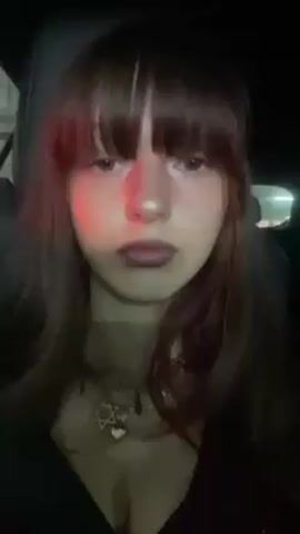 A young woman with brown hair and bangs, wearing a dark top and a necklace with a Star of David pendant, is sitting in what appears to be a car at night. The lighting is dim, and she looks directly at the camera. She briefly raises her hand to her hair.