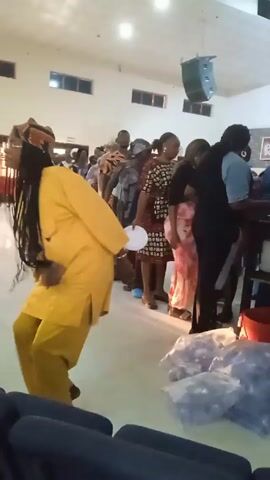 The video shows a line of people waiting to receive food in what appears to be a church hall. A woman in a yellow outfit dances while holding a white plate. Other people in the line are dressed in various clothes. There are bags of what appears to be drinks stacked near some chairs, and a speaker hangs from the ceiling.
