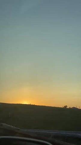 The video shows a sunset landscape viewed through the window of a moving vehicle. The sky is a gradient of light blue at the top, transitioning to orange and yellow near the horizon. The sun is setting behind a dark, grassy hill. A series of gray pipelines runs across the hillside, partially obscuring the view of the sunset. A small signpost is visible on the hill. The side mirror of the vehicle is visible in the lower left corner of the frame. Music plays in the background.