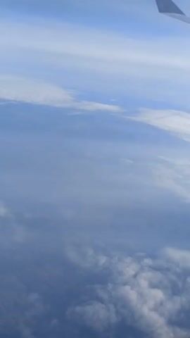 The video depicts a view from an airplane window, showcasing the sky and clouds. A portion of the airplane wing is visible in the upper right corner of the frame. The sky is light blue, with varying layers of clouds, some appearing thin and wispy, and others denser and lower in the frame. The overall scene is serene and typical of a view during air travel.