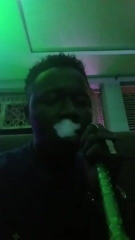 A person is sitting in a dark room with dynamic colorful lighting, inhaling from a green and silver hookah and exhaling a cloud of white smoke. The person has curly hair. The background includes a wall with geometric patterns and a window with partially closed blinds. The lighting shifts between green and blue with hints of purple.