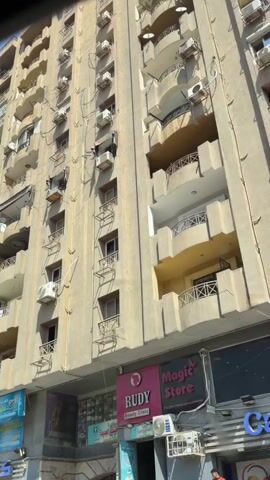 The video pans upward across the facade of a multi-story apartment building, showcasing numerous balconies and air conditioning units. The building is constructed of beige concrete. A blue sign with Arabic text is visible on the lower part of the building, along with other store signs, including "RUDY Beauty Chain" and "Magic Store." To the right, a portion of a yellow building is visible. The sky is clear and blue. The camera then pans to the left, revealing more apartment buildings and lush green trees. In the foreground, the rear of a silver car is visible, and to the left, a tuk-tuk with passengers is driving past. A man wearing a white t-shirt and dark pants stands near the sidewalk in the lower left quadrant of the frame. The scene appears to be a street view in a residential urban area.