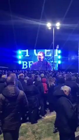 A large crowd of people is gathered outside in front of a large stage and screen. The screen displays an image of a man and text. The stage is illuminated with bright lights, and people can be seen standing on the stage. The crowd is facing the stage, presumably watching a presentation or performance. The environment appears to be urban, with streetlights visible. The video is shot from within the crowd, providing a view of the event from the audience's perspective. The people in the crowd are dressed in casual clothing, appropriate for an outdoor gathering. The lighting suggests it is nighttime or dusk. The overall atmosphere appears to be that of a public event or rally.