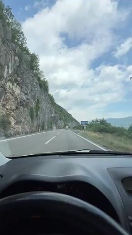 The video shows a scenic drive through a mountainous region in Frutak, Montenegro. The first shot is from the driver's perspective, looking forward through the windshield at a winding road. A steep, rocky mountain covered with some vegetation borders the left side of the road, while the right side features lower green hills and distant mountains. A large billboard displaying "BIANCA" is visible on the right. The sky is bright with scattered clouds, indicating daytime. The car's dashboard and steering wheel are in the foreground. The second shot focuses on the car's interior, showing a woman in the passenger seat. She has long blonde hair, wears a white shirt and a black baseball cap, and is holding a smartphone with both hands, recording the landscape outside the car window.