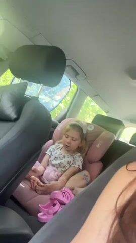 A young girl with blonde hair and a pink bow is sitting in a car seat in the back seat of a car. She is wearing a white shirt with a floral pattern and a pink skirt. She has pink sandals on the floor in front of her. The car interior is visible, with leather seats and a sunshade on the window. The girl appears to be talking or singing.