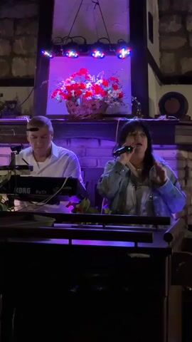A man and a woman are performing music on stage. The man is playing a Korg Pa5X keyboard. The woman is singing into a microphone. The stage is decorated with a floral arrangement. The lighting changes color throughout the video, cycling through blue, red, and purple. The background appears to be a stone wall with a fireplace.