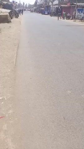 The video shows a road in a town. The road is paved. There are people, structures, and vehicles visible in the distance. One side of the road has a dirt shoulder, while the other appears to be lined with shops. The camera moves along the road.