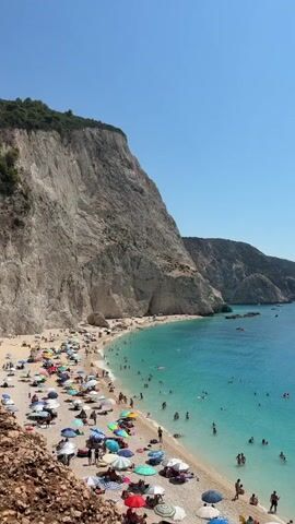 The video shows a crowded beach with white pebbles and turquoise water. Steep, rocky cliffs rise behind the beach. Many people are sunbathing under colorful umbrellas, and others are swimming and playing in the clear water. A large white yacht is anchored in the bay, and there is a small sailboat in the distance. The sky is a clear, bright blue, indicating a sunny day.