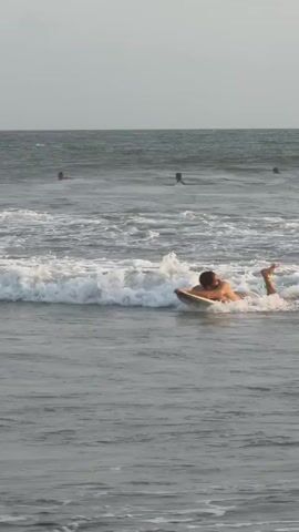 The video shows several surfers in the ocean. In the foreground, a man is lying on a surfboard, paddling through a wave. He is wearing swim trunks and has short, dark hair. In the background, several other surfers are visible, some lying on their boards and others paddling. The ocean is choppy, with white waves breaking. The sky is overcast.