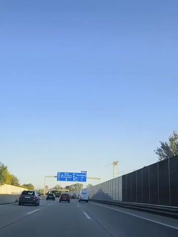 A highway scene with a clear blue sky. Multiple cars are traveling on the road. A large blue directional sign is visible overhead, indicating directions to Wien, Floridsdorf, ASt Strebersdorf, and Korneuburg. The sign also shows route numbers A22 and 16. On the right side of the highway, there is a tall sound barrier. A construction crane is visible in the background. The camera is positioned inside a vehicle, suggesting it is recording from a driver's or passenger's perspective.