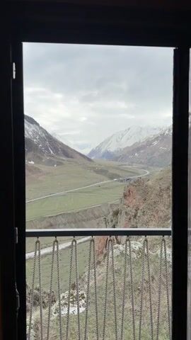 The video presents a scenic view of a mountain valley, seen through the frame of a window and balcony railing. The foreground features a balcony railing made of dark metal, with rope-like details suspended below the rail. The view beyond reveals a wide valley surrounded by mountains, many capped with snow. A river runs along the valley floor, and a highway cuts through the landscape. The sky is overcast with a blanket of grey clouds. The camera slowly zooms in and tilts slightly upward to better capture the valley scene.