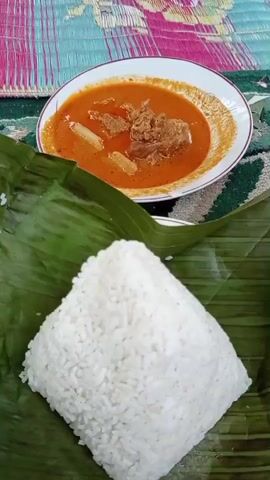 A plate of curry is in the background, and a pile of rice wrapped in banana leaves is in the foreground. The food is placed on a mat with a colorful striped pattern.