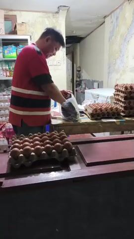 A man wearing a red shirt with black and white stripes is standing behind a wooden counter. He is holding a clear plastic bag with dark items inside and appears to be placing it down on the counter. In front of him are two rows of egg cartons filled with brown eggs. To his right, stacked higher, are more egg cartons. In the background, shelves stocked with various items and a white refrigerator are visible. The walls in the background are textured and appear worn. The lighting is dim.