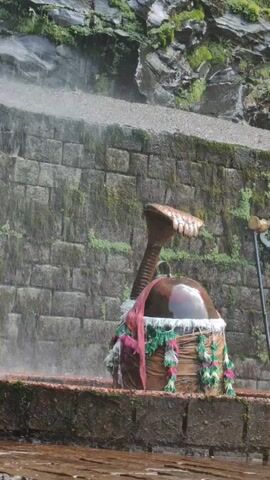 A statue of Shiva adorned with a cobra hood is shown partially obscured by a wall of stacked, moss-covered stones. Water cascades down the wall and onto the statue, which is also wrapped in fabric and garlands. The ground in front of the statue is wet and appears to be made of wood. The scene is outdoors with greenery visible at the top of the frame.
