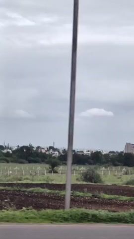 The video shows a panning shot of a rural landscape under a cloudy sky. In the foreground, there is a plowed field with some green vegetation. In the midground, there is a fence with posts and some trees. In the background, there is a town with buildings and some trees. The sky is overcast with gray clouds.