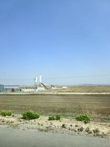 The video shows a landscape with a clear blue sky. In the distance, there is an industrial facility with buildings and silos. The foreground consists of dry, patchy fields and a road. The camera appears to be moving, likely from a vehicle, as evidenced by the slight blur and the perspective. The overall impression is of a rural or semi-industrial area under a bright sky.