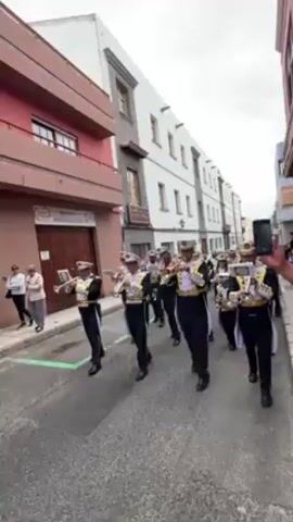 The video shows a brass band marching down a city street on what appears to be an overcast day. The band members are wearing distinctive uniforms: black pants and jackets with gold and white accents, along with white hats. They are playing brass instruments, including trumpets and trombones. Several pedestrians are standing or walking on the sidewalks, observing the band as it passes. Buildings of various colors and architectural styles line the street. A person holding a smartphone can also be seen, suggesting they are recording the performance.