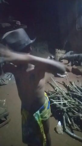 The video shows a young boy drinking from a silver pot. He is wearing shorts and no shirt. There are other people around him, but they are not in focus. The video appears to be taken outdoors at night. There is firewood on the ground.