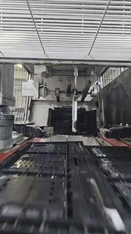 The video shows a large industrial machine with moving parts. A cutting tool is visible and appears to be in operation. The machine is enclosed in a metallic structure with a slatted ceiling. The environment seems to be a factory or workshop. There are no people visible in the video.