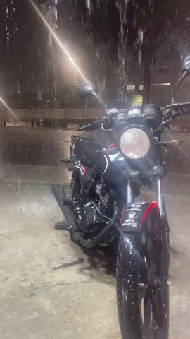 The video shows a black motorcycle parked outdoors at night. Rain is falling heavily, creating a wet environment with water splashing on the ground and the motorcycle. The lighting suggests an urban setting, possibly a street or a parking area, with artificial lights visible in the background. The focus is on the motorcycle, highlighting its details and the effect of the rain on its surfaces. There are no people visible in the video.