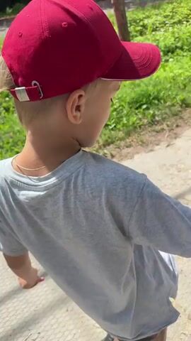 A boy wearing a red baseball cap and a grey t-shirt with a graphic print stands in an outdoor environment. He turns and faces the camera, opening his mouth as if to speak or sing. He has blond hair, visible from beneath his cap. The background consists of green foliage and a path.