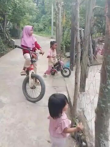A child in a pink shirt and striped pants stands on a concrete path, facing away from the camera and towards a wire fence. Behind the child, two other children are on bicycles. One child, wearing a pink hijab and red shirt, is on a red and white bicycle. The other child, in a pink shirt, is on a smaller bicycle. To the right of the path, there is a yard with a house and several bicycles.