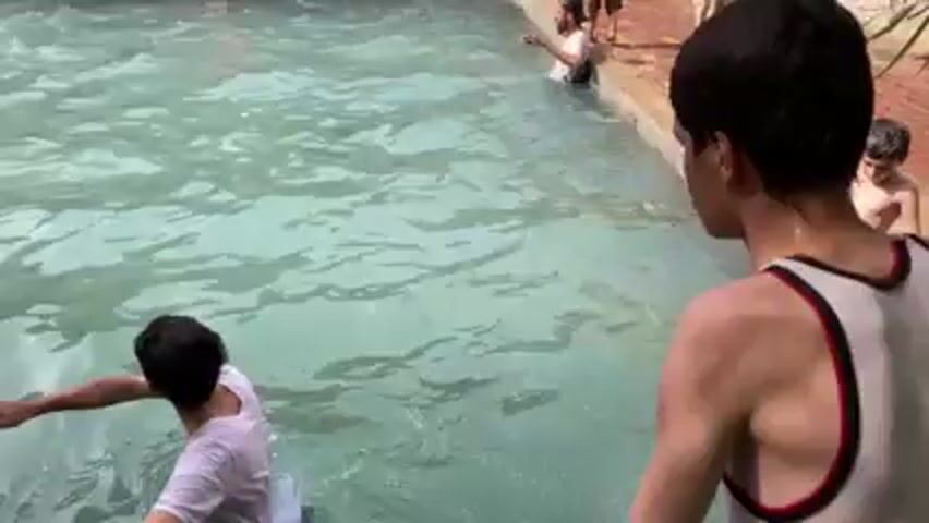 The video shows a person in a white tank top and dark shorts doing a backwards flip into a body of water. Other people are standing and sitting on the edge of the pool, and wading in the water. The water appears to be a milky blue-green. The edge of the pool is made of red brick.