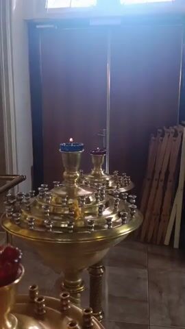 The video shows a close-up of several brass candelabras with lit candles. The candelabras are large and ornate, with multiple holders for candles arranged in tiers. Some of the candles are in small, colored glass cups. In the background, there is a wooden door and a stack of wooden chairs. The lighting is warm and appears to be coming from above, casting a glow on the brass. The overall scene suggests an indoor religious setting, likely a church or chapel.