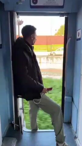 A young man stands inside a blue-toned train car. The train door is open, and the man leans against the doorframe as the train moves past a street with a few buildings and some shrubbery. The man has on a black jacket and light green pants, and he is using his cell phone.