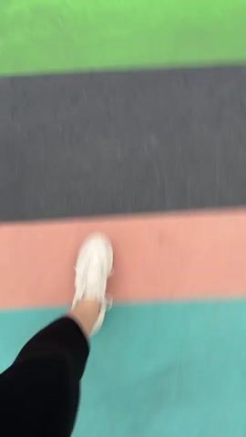 A first-person perspective video shows a person walking across a striped crosswalk. The crosswalk is composed of horizontal stripes of yellow, green, grey, and pink. The person is wearing white sneakers, black leggings, and a dark blue shirt. The video is filmed from the perspective of the person's legs and feet, capturing their movement. The lighting is neutral, and the environment appears to be outdoors.