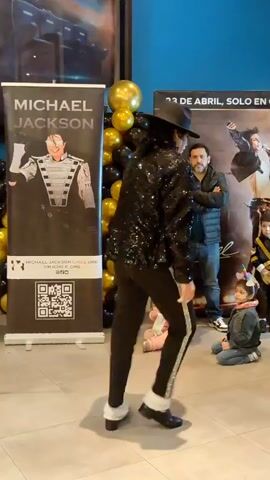 The video shows a Michael Jackson impersonator standing and looking away from the camera. The impersonator is wearing a black sequined jacket, black pants with a silver stripe down the side, black shoes, and a black fedora. The impersonator is standing in front of a banner that says "MICHAEL JACKSON" with a picture of Michael Jackson in a military-style uniform. To the right of the banner, there is a balloon arch made of black and gold balloons. In the background, there is another banner with Spanish text that reads "23 DE ABRIL, SOLO EN CINE". There are several children in the background, one of whom is wearing a unicorn headband. The floor is tiled. The lighting appears to be artificial. The video is shaky and appears to be taken by someone holding a phone.