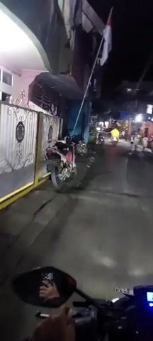 This video shows a street scene at night. A person is riding a motorcycle, with the camera mounted on the handlebars. To the left, there is a gate and a building. To the right, there are more buildings and several parked motorcycles. Further down the street, there are people walking and more motorcycles. The street is illuminated by artificial lights.