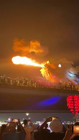 The video captures a dramatic nighttime scene featuring a large, illuminated dragon sculpture on a bridge, breathing fire. The dragon's head is predominantly yellow and green, and it projects a sizable burst of flames into the dark sky, leaving a trail of smoke. A crowd of spectators lines the bridge behind the dragon, many holding up their phones to record the spectacle. The foreground shows more people, also with phones in hand, recording the event. A large, heart-shaped decoration made of red lights is also visible in the foreground, slightly off to the side. The overall setting appears to be an urban waterfront at night, enhanced by artificial lighting and the spectacle of the fire-breathing dragon.
