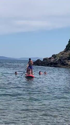 A group of people are in the water near a rocky shore. One person is standing on a paddleboard, holding a paddle. Two other people are sitting on the paddleboard behind the person standing. Two more people are in the water swimming or wading. The water is calm and clear. The sky is blue with a few clouds. There is a rocky island with green vegetation in the background.