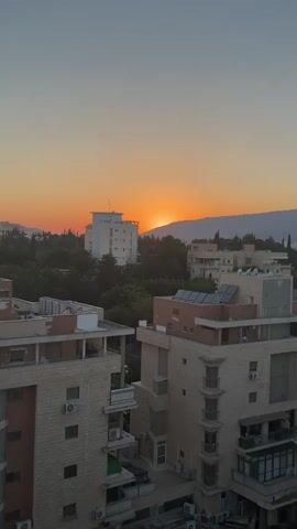 The video is a vertical shot of a sunset over a cityscape. The sun is partially obscured by a mountain range in the distance, casting an orange glow across the sky. In the foreground, there are several buildings, including apartment complexes with balconies and rooftop solar panels. Trees are visible between the buildings, and the overall impression is one of a tranquil evening scene. There are no people or animals visible in the video.