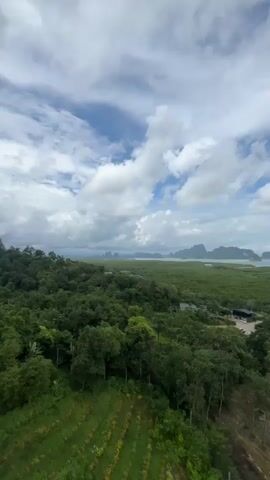 A drone shot pans across a lush green landscape under a cloudy sky. In the foreground, terraced fields of crops stretch across a hillside. Further back, a dense forest covers rolling terrain, giving way to a vast expanse of mangrove trees that reach toward the sea. Several islands with steep, jungle-covered slopes rise from the water in the distance, their peaks partially obscured by clouds. A few buildings and a cellular tower are visible amidst the trees. The overall scene depicts a tropical coastal environment with abundant vegetation and striking geological formations.