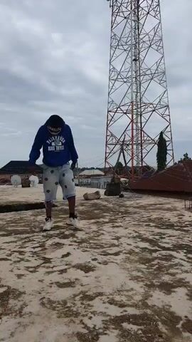This media file captures a young man standing on a rooftop in Anguldi, Nigeria. He is wearing a blue hoodie with the words "FAIRFIELD BLUE DEVILS" visible on it, along with light-colored denim shorts and white sneakers. He also has on white goggles or protective eyewear and a face mask. The man is positioned centrally in the frame, with a large, red and silver transmission tower dominating the background. The sky is overcast, suggesting a cloudy or possibly rainy day.

The man appears to be engaged in some form of activity, possibly dancing or posing, as his body language changes slightly throughout the short clip. In the first few seconds, he bends his head down and has his hands by his sides. Then, he raises his hands and strikes a pose, with one hand on his hip and the other raised. His stance is dynamic, and he seems focused on the action.

The setting is an urban or semi-urban environment, indicated by the presence of buildings with satellite dishes and the prominent telecommunications tower. The rooftop itself is concrete and shows signs of wear and tear, with patches of discoloration. The overall atmosphere is somewhat subdued due to the cloudy weather. There are no other people visibly present in the frame. The time of day is indeterminate due to the overcast conditions, but it appears to be daytime.
