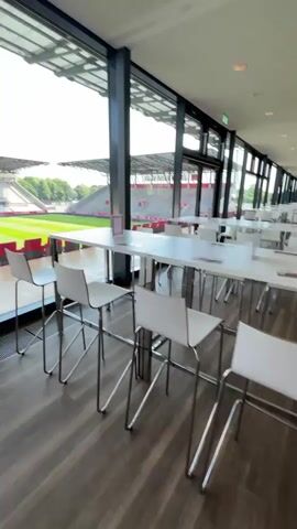 The video shows a view of a soccer stadium from inside an enclosed room with large windows. The camera slowly pans from left to right, focusing on the stadium view. The stadium has a green field, red seats, and a checkered pattern in the stands. The sky is blue with a few clouds. Inside the room are white tables and chairs, suggesting a lounge or dining area. The room has wood floors and a metallic grate near the window. The windows are large and provide a clear view of the stadium.