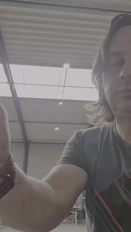 The video begins with a close-up, shoulder-level shot of a man's right arm and forearm. He is wearing a dark t-shirt with a graphic design on the right side of his chest and a watch with a reddish-brown band on his left wrist. His arm is flexed, and his hand is out of frame at the top left. The background is a brightly lit industrial-looking space with a corrugated ceiling and visible light fixtures. As the video progresses, the man's face comes into partial view on the right side of the frame. He has medium-length brown hair and is looking towards the camera with a neutral expression. The arm continues to move, and in the final seconds, the man smiles slightly.