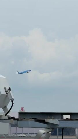 A blue airplane ascends into a cloudy sky, seen from below and to the side, with the edge of a large, industrial-looking piece of equipment dominating the foreground. The airplane is positioned in the upper left quadrant of the frame and is angled slightly upward, indicating it is in the process of takeoff or climbing. The sky is overcast with soft, white clouds against a pale blue background. The foreground equipment appears to be part of an airport structure, possibly a gantry or a building's exterior, with visible metal beams, a large cylindrical object, and cables. The lighting suggests it is daytime.