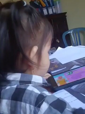 A toddler, seen from behind and to the side, sits at a desk wearing a black and white checkered shirt. The child is looking at a smartphone placed on the desk. The phone screen displays a colorful animation, possibly a cartoon or educational game. Several papers and books are also on the desk, and a dark wooden cabinet is visible in the background, along with a blue chair. The child's dark hair is pulled back in a ponytail.