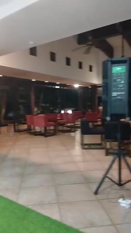 The camera pans across a hotel lobby or common area. The flooring is square tiles, and there are multiple seating arrangements consisting of dark wood tables and red upholstered chairs. A black speaker system on a tripod stands near a couch with red floral pattern. The exterior wall is largely open, offering a view of the night. Music is playing.