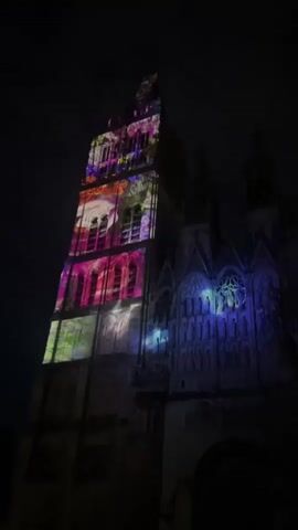 A large stone building is illuminated at night with colorful light projections. The projections show various floral and abstract patterns, primarily in shades of pink, purple, green, and yellow, covering a tall tower and parts of the adjacent structure. The sky above is dark.