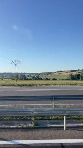 The video shows a view from a moving vehicle, likely a car, looking out over a rural landscape. The foreground features a metallic guardrail. Beyond the guardrail, a paved road is visible, with a white line marking its edge. Further in the distance, agricultural fields are apparent, with some appearing to be crops and others possibly meadows. A single tall electrical pylon with wires extending from it stands in the middle ground. The sky is a clear, bright blue, with no clouds visible. The overall environment suggests a daytime scene in a countryside setting, possibly during the warmer months given the greenery. There are no people or animals visible in the frame.