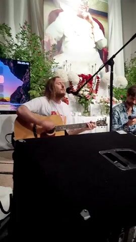 A group of four men are performing music on a stage. The man on the far left is singing and playing an acoustic guitar. He is wearing a white t-shirt. To his right, another man is looking at his phone and wearing a patterned shirt. In the center, a man is playing a red bass guitar and wearing a white t-shirt with a graphic. To his right, a man wearing a black shirt is singing into a microphone and sitting on the floor. Behind the performers, a large portrait of a person is displayed on a white backdrop, surrounded by floral arrangements and greenery. A microphone stand is positioned in front of the man playing the acoustic guitar. A black speaker is in the foreground, obscuring part of the stage. Another acoustic guitar lies on the floor in the lower right corner of the frame. The lighting is soft and warm.